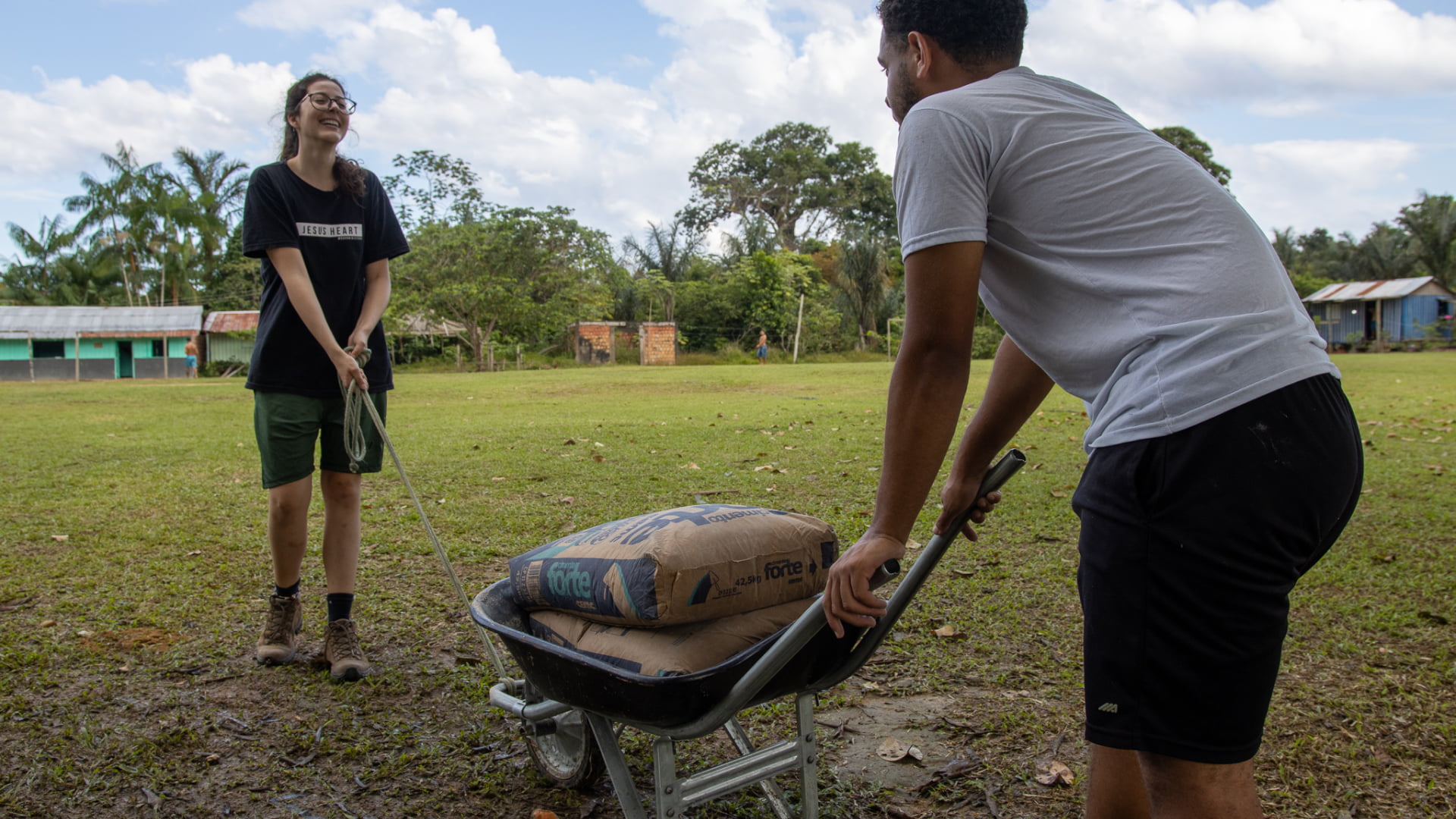 Dois jovens se ajudando, carregando um carrinho com sacos de cimento