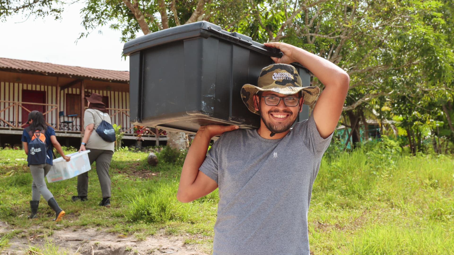 Rapaz sorrindo carregando uma caixa nos ombros com um chapéu do Missões Noroeste